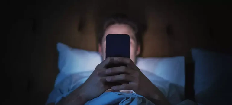 Um homem deita na cama e observa o celular.