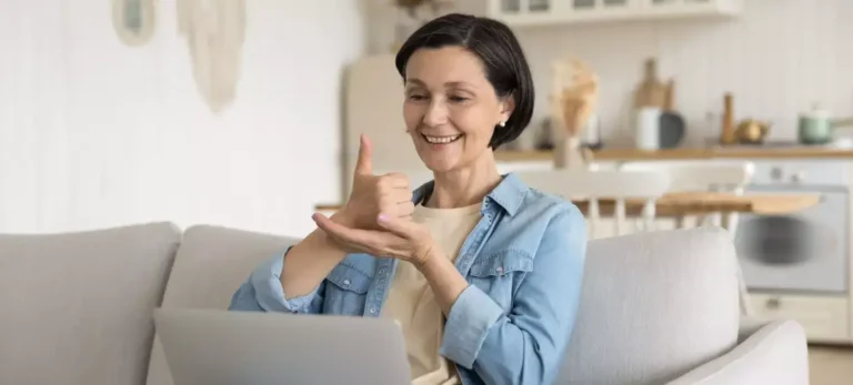 Uma mulher de cabelos curtos e castanhos faz um sinal de Libras para uma tela de computador, à sua frente.