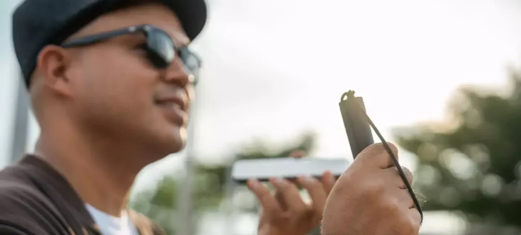 Um homem com deficiência visual usa um boné, óculos escuros e em uma mão segura uma bengala. Na outra, segura um smartphone.