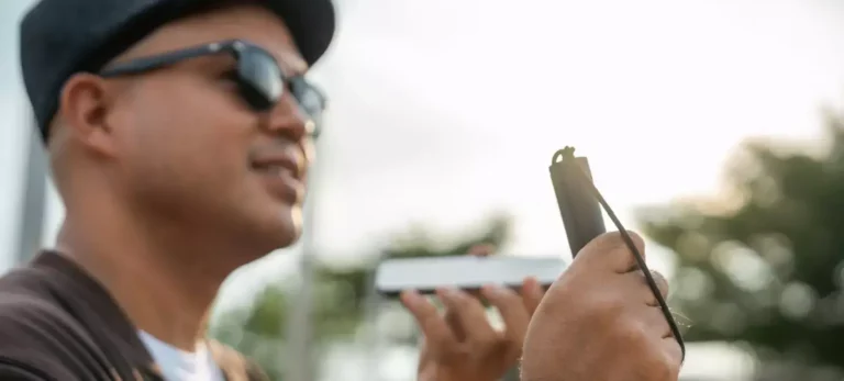 Um homem com deficiência visual usa um boné, óculos escuros e em uma mão segura uma bengala. Na outra, segura um smartphone.