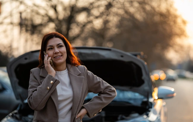 Mulher fala ao celular ao lado de um carro com o capô aberto, em uma rua, enquanto aguarda assistência.